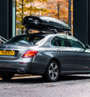 Grey Mercedes-Benz E-Class parked on an urban street with a black roof box for extra storage, ready for a comfortable city transfer.