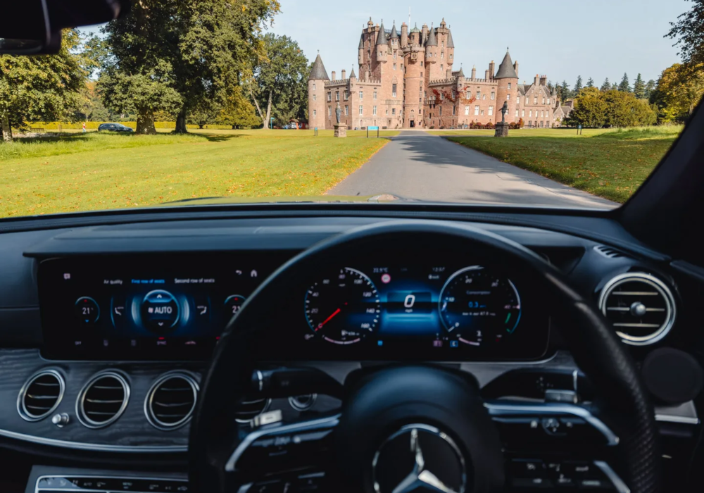View from inside a Mercedes-Benz showcasing its advanced dashboard, with Glamis Castle standing majestically in the Scottish countryside ahead.