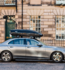 A grey Mercedes-Benz E-Class with a roof box parked on a cobbled street in front of historical buildings.